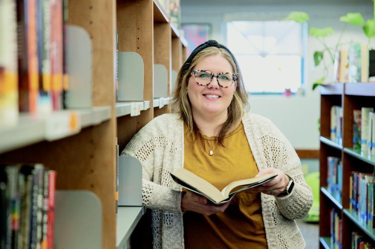 A smiling woman stands in a library, holding an open book. She is wearing glasses, a mustard-colored shirt, and a cream knit cardigan, with bookshelves on either side and soft natural light coming through a window in the background.