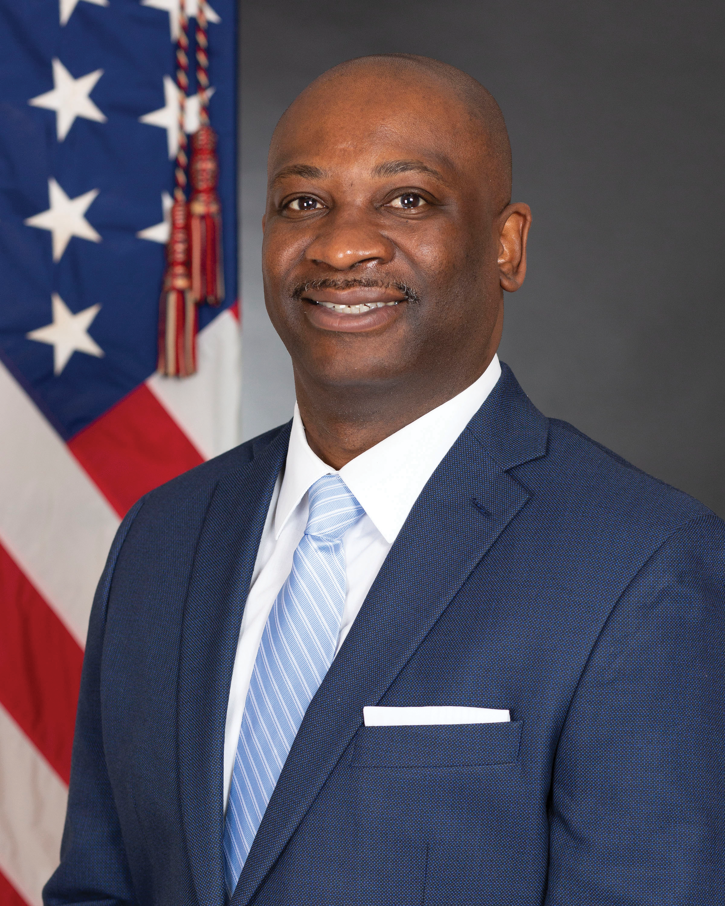 The image shows a man in formal attire, wearing a blue suit with a light blue striped tie and white shirt. He is smiling, standing in front of a U.S. flag. The background is a solid dark gray.